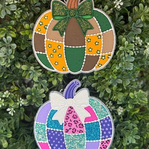 May include: Two decorative pumpkin ornaments featuring patchwork designs. The top pumpkin has orange, green, and brown segments with a glittery green bow. The bottom pumpkin features purple, pink, and teal segments with a white glittery bow.