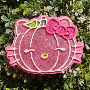 May include: Pink, glittery air freshener in the shape of a Hello Kitty pumpkin. The design features a pink bow, white outlines, and a green stem. The air freshener has a small, yellow nose and closed eyes. The text "little freshie shop" is visible.
