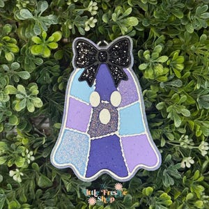 May include: A ghost-shaped air freshener featuring a patchwork design of light blue, lavender, and dark purple sections with glittery textures. A black glittery bow is attached to the top. The text Little Freshie Shop is printed at the bottom.
