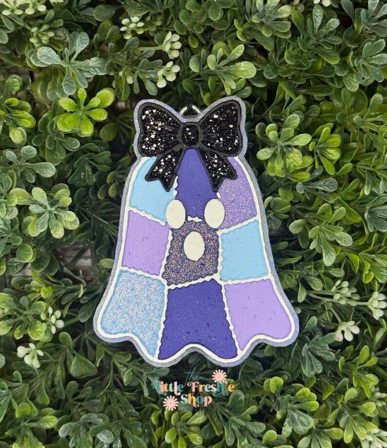 Peut inclure: Un d&eacute;sodorisant en forme de fant&ocirc;me avec un motif en patchwork compos&eacute; de sections bleu clair, lavande et violet fonc&eacute; aux textures paillet&eacute;es. Un n&oelig;ud noir paillet&eacute; est fix&eacute; au sommet. Le texte Little Freshie Shop est inscrit en bas.