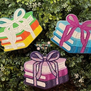May include: Three colorful book-shaped ornaments with white bows. The books are stacked and tied with a ribbon. The books are in shades of green, orange, yellow, blue, and purple. The ornaments are on a green leafy background.