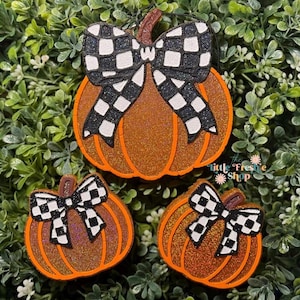 May include: Three glittery orange pumpkin decorations with black and white checkered bows. The pumpkins have brown stems and are set against a backdrop of green foliage. The text "the little freshie shop" is visible.