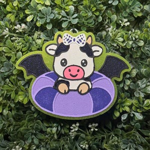 May include: A glittery ornament featuring a black and white cow with a pink snout and a white bow, sitting in a purple and blue inner tube. The cow has black bat wings and is framed by a bright lime green border with a silver hanging loop.