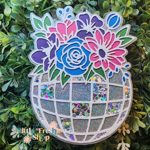 May include: A decorative ornament featuring a floral arrangement in a globe-shaped container. The flowers are in shades of blue, pink, and purple, with green leaves. The globe has a glittery, silver finish. The ornament has the text "littl Fresh Shop".