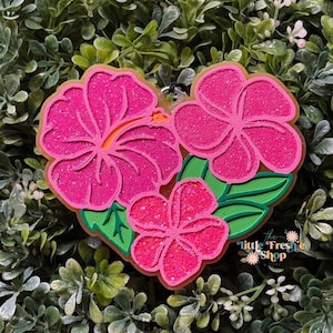 May include: A heart-shaped keychain featuring three pink hibiscus flowers with green leaves. The flowers have glitter accents and orange stamens. The text "the little freshie shop" is visible.