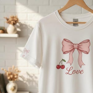 May include: White t-shirt with a pink bow and cherry graphic, plus the word "Love" in script. A small bow and the letter "C" are on the left sleeve. The tee is displayed on a wooden hanger.