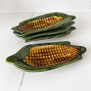 May include: A collection of ceramic corn-shaped serving dishes. The dishes are green with a textured design on the outer edges, and the inner portion is a golden-brown colour, mimicking the appearance of corn kernels. The dishes are arranged on a white surface.