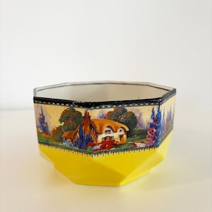 May include: A hexagonal ceramic bowl with a yellow base and a black rim. The sides feature a landscape scene with a cottage, trees, and flowers. The bowl is likely vintage and decorative.