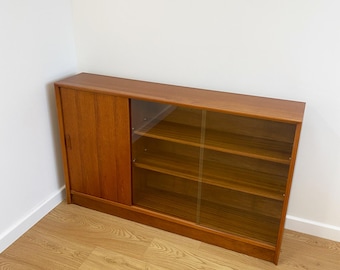 May include: A mid-century modern wooden cabinet with a sliding door and glass-fronted shelves. The cabinet is made of warm-toned wood and has a rectangular shape. The glass shelves are visible through the glass door.