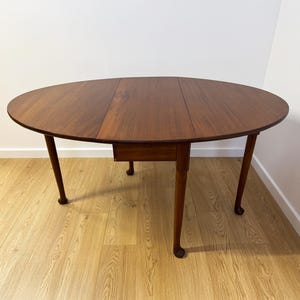 May include: A dark brown, oval wooden table with a drop-leaf design. The table has four slender, curved legs and a small drawer. The table is made of wood and has a smooth, polished surface.