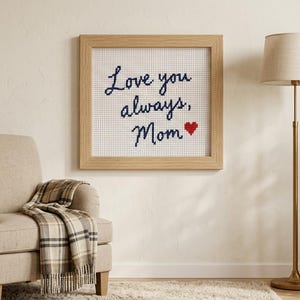 May include: A square framed cross-stitch artwork featuring the dark blue embroidered text "Love you always, Mom" followed by a small red heart, displayed on a white grid fabric within a light-colored wooden frame.