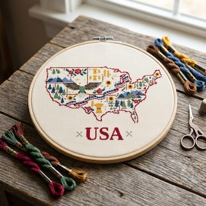 May include: A circular wooden embroidery hoop containing a cross-stitch map of the USA. The map features a central bald eagle, mountains, trees, flowers, and wheat stalks. The text USA is stitched in red below the map, flanked by small green crosses.