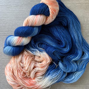 May include: A skein of yarn featuring a gradient of deep navy blue, light blue, and soft peach tones. The yarn is twisted into a compact bundle at the top, while the bottom section is loose and flowing, showcasing the blended colour transitions.