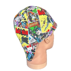 May include: A colorful, patterned cap featuring comic book panels with the Batman logo and characters. The cap is made of fabric and has a curved brim. The design includes text such as "Batman" and "Detective".