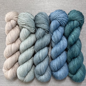 Könnte beinhalten: Fünf Stränge Garn in einer Reihe, jeder in einem anderen Blau- und Grünton. Die Farben reichen von hellem Beige bis zu tiefem Teal. Das Garn ist fest gewickelt und wirkt weich.