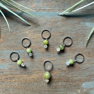 Peut inclure: Un ensemble de six marqueurs de mailles, chacun composé d'un anneau en métal foncé, d'une perle ronde vert olive et d'une petite perle en cristal transparent à facettes suspendue. Les marqueurs sont disposés en cercle sur une surface en bois.