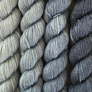 May include: Six skeins of twisted yarn arranged in a vertical gradient, transitioning from a light cream and pale silver-grey on the left to medium grey, slate blue, dark charcoal, and deep black on the right, showcasing a soft, fibrous texture.