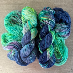 Puede incluir: Dos madejas de hilo colorido en tonos azules, verdes y morados. El hilo está dispuesto en una madeja torcida y una madeja suelta. El hilo tiene un patrón de color abigarrado, con azules y verdes más oscuros que pasan a tonos más claros.
