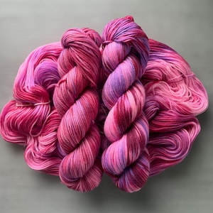 Puede incluir: Primer plano de varios ovillos de hilo teñido a mano en tonos rosa, magenta y morado. El hilo está enrollado firmemente en grandes madejas texturizadas, con algunos hilos sueltos visibles. Las transiciones de color crean un efecto degradado. El fondo es gris liso.