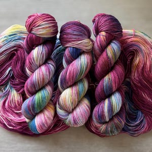 May include: Three skeins of hand-dyed yarn in a vibrant array of colors. The yarn is a mix of deep magenta, blues, greens, yellows, and white. The skeins are twisted into a rope-like shape, with the ends of the yarn fanned out.