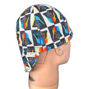 Puede incluir: Una gorra de soldador con un patrón de cuadros blancos y negros y un gráfico colorido repetitivo de un personaje con casco. La gorra incluye una pequeña etiqueta de tela blanca que dice Bo-Beanies Made in Canada en el borde posterior inferior.