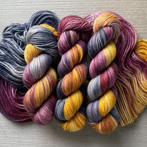 May include: A collection of colorful yarn skeins. The yarn features a gradient of colors, including shades of purple, yellow, orange, and gray. The skeins are arranged on a light gray wooden surface, showcasing the texture and hues of the fiber.