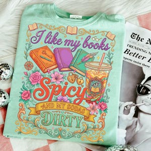 May include: A light green t-shirt with a colorful graphic design. The design includes the text "I like my books Spicy and my sodas Dirty" with illustrations of books, flowers, and a drink. The shirt is folded on a white surface.