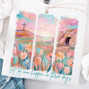 Op de afbeelding: Wit sweatshirt met lange mouwen met een kleurrijke grafische print van een landschap met een kruis, bloemen en een graf. De tekst "A lot can happen in three days" staat eronder. Het sweatshirt wordt getoond met een blauwe jeans.