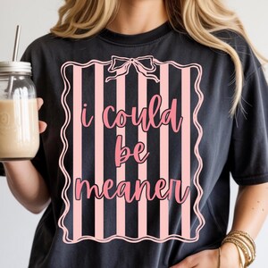 May include: Black t-shirt with the text "I could be meaner" in pink script inside a pink and white striped frame. The shirt is worn with denim shorts and gold bracelets. A glass jar with a straw and a beige drink is held in the left hand.
