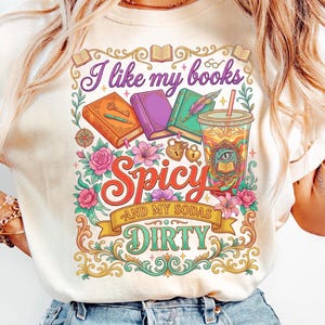 May include: Cream-colored t-shirt with a colorful graphic design. The design features the text "I like my books Spicy and my sodas Dirty" with illustrations of books, flowers, and a drink. The shirt has short sleeves and a crew neck.