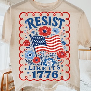 Può includere: Una t-shirt color crema con un design grafico rosso, bianco e blu. La stampa include una bandiera americana centrale circondata da motivi floreali, racchiusa in un bordo smerlato rosso, con il testo RESIST LIKE IT'S 1776 in blu.