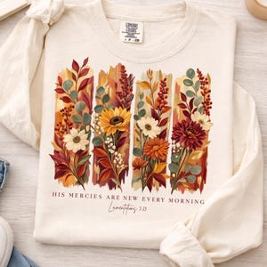 May include: A cream-colored long-sleeve shirt featuring a graphic of four vertical brushstrokes filled with autumn-toned flowers and foliage. Below the floral design, the text reads, "HIS MERCIES ARE NEW EVERY MORNING" followed by "Lamentations 3:23".