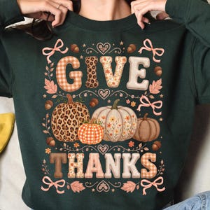 Op de afbeelding: Een donkergroen sweatshirt met lange mouwen en de tekst GIVE THANKS in grote blokletters met diverse patronen. Het ontwerp bevat illustraties van drie pompoenen, eikels, bladeren en roze strikken in een decoratieve opstelling.