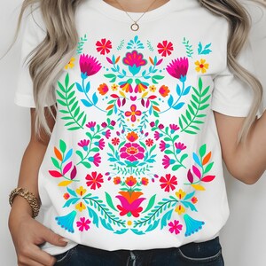 May include: White t-shirt featuring a vibrant floral design. The design includes pink, blue, yellow, and orange flowers with green leaves. The shirt has a crew neck and short sleeves, perfect for casual wear.