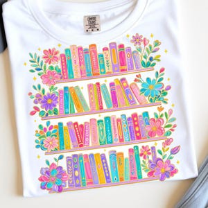 Può includere: T-shirt bianco con una colorata illustrazione di una libreria con libri e fiori. Il design presenta dorsi di libri in varie tonalità, tra cui rosa, blu e giallo. La maglietta ha un girocollo e maniche corte.