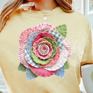 May include: A yellow t-shirt features a large, colorful fabric rose applique. The rose is made of various patterned fabrics in shades of pink, blue, green, and white, with green fabric leaves. The shirt has a relaxed fit and short sleeves.