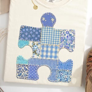 Puede incluir: Camiseta color crema con un gran diseño de pieza de rompecabezas de patchwork en varios tonos de azul. La pieza del rompecabezas está hecha de diferentes telas estampadas, incluyendo floral, cuadros y azul liso. La camiseta tiene cuello redondo y mangas cortas.
