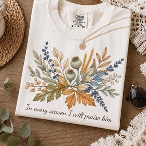 May include: An off-white short-sleeve t-shirt featuring a central botanical illustration of autumn leaves, berries, and seed pods. Below the graphic, the text reads, In every season I will praise him. The shirt has a crew neckline and a Comfort Colors label.