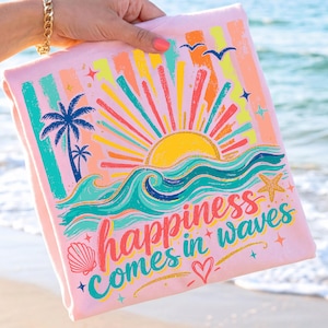 Op de afbeelding: Een roze t-shirt met een kleurrijke strandafbeelding. Het ontwerp bevat een gele zon, blauwe golven, palmbomen en de tekst "happiness comes in waves". Het shirt wordt door een persoon op het strand omhoog gehouden.
