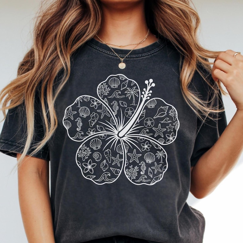 May include: A dark gray t-shirt featuring a large white floral design filled with seashells, starfish, seahorses, and palm trees. The design is centered on the front of the shirt, creating a beach or tropical theme.