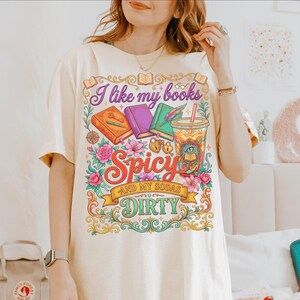 May include: Cream-colored t-shirt with a colorful graphic. The design features the text "I like my books Spicy and my sodas Dirty" with illustrations of books, flowers, and a drink. The shirt has short sleeves.