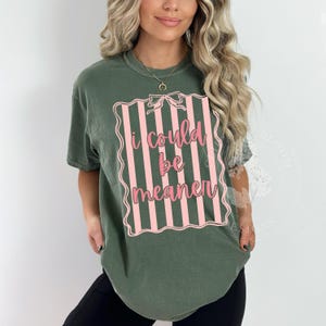 May include: Olive green t-shirt with the text "I could be meaner" in pink script within a pink and white striped frame. The shirt has short sleeves and a crew neck.