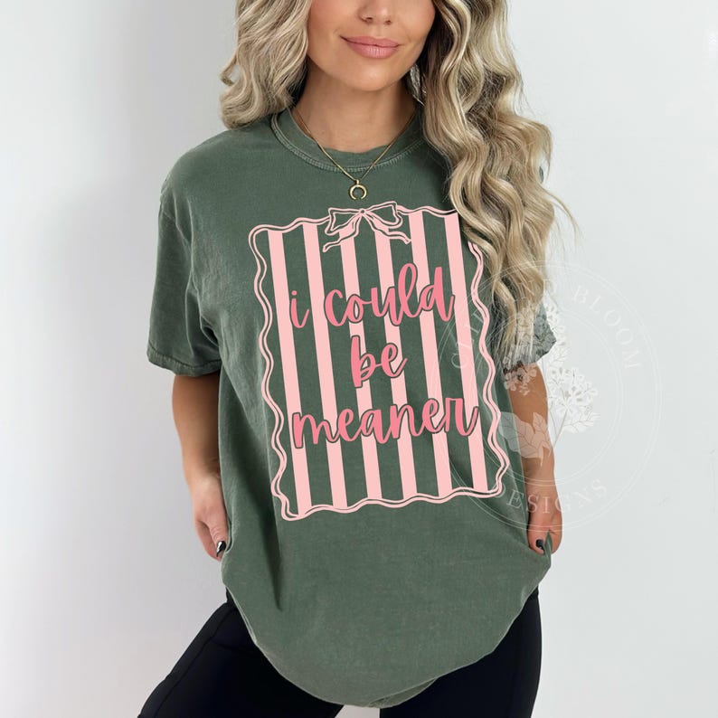 May include: Olive green t-shirt with the text "I could be meaner" in pink script within a pink and white striped frame. The shirt has short sleeves and a crew neck.
