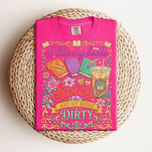 May include: A hot pink t-shirt with a colorful graphic design. The design includes the text "I like my books Spicy and my sodas Dirty" along with illustrations of books, flowers, and a drink. The shirt is folded on a woven surface.