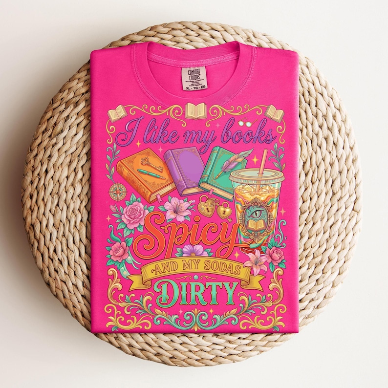 May include: A hot pink t-shirt with a colorful graphic design. The design includes the text "I like my books Spicy and my sodas Dirty" along with illustrations of books, flowers, and a drink. The shirt is folded on a woven surface.