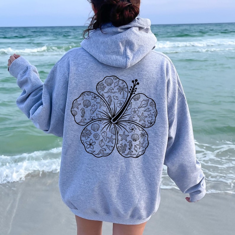 May include: A light gray hoodie with a large black floral design on the back. The floral design is filled with various sea-themed illustrations, including seashells, starfish, and seahorses. The hoodie has a hood and long sleeves.