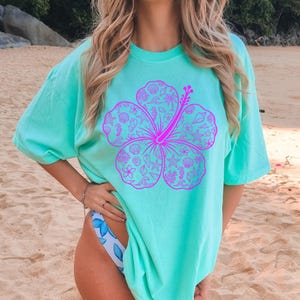 May include: A turquoise t-shirt featuring a large, vibrant pink hibiscus flower design. The flower is filled with seashell and tropical elements. The shirt has short sleeves and a relaxed fit, perfect for a beach day.