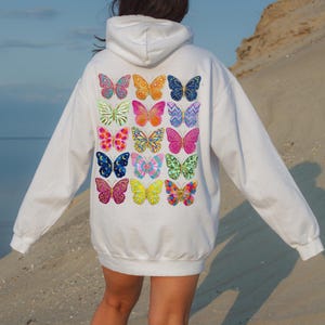 May include: White hoodie featuring a colorful butterfly design on the back. The butterflies are in various colors and patterns, including pink, blue, orange, and yellow. The hoodie has a hood and long sleeves.