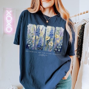 Puede incluir: Camiseta azul marino con un dise&ntilde;o floral con flores amarillas, azules y moradas. La camiseta tiene el texto "HIS MERCIES ARE NEW EVERY MORNING" y "Lamentations 3:23". La camiseta es de estilo casual.