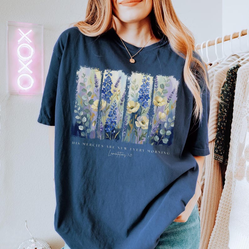 Puede incluir: Camiseta azul marino con un dise&ntilde;o floral con flores amarillas, azules y moradas. La camiseta tiene el texto "HIS MERCIES ARE NEW EVERY MORNING" y "Lamentations 3:23". La camiseta es de estilo casual.